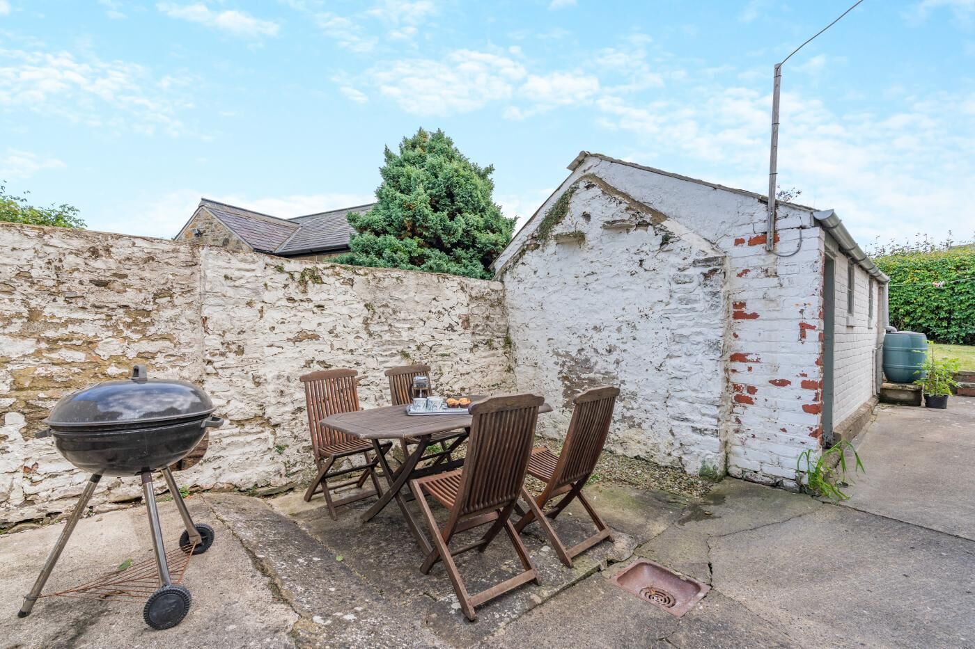 Een terras buiten met een barbecue, een houten tafel en stoelen.