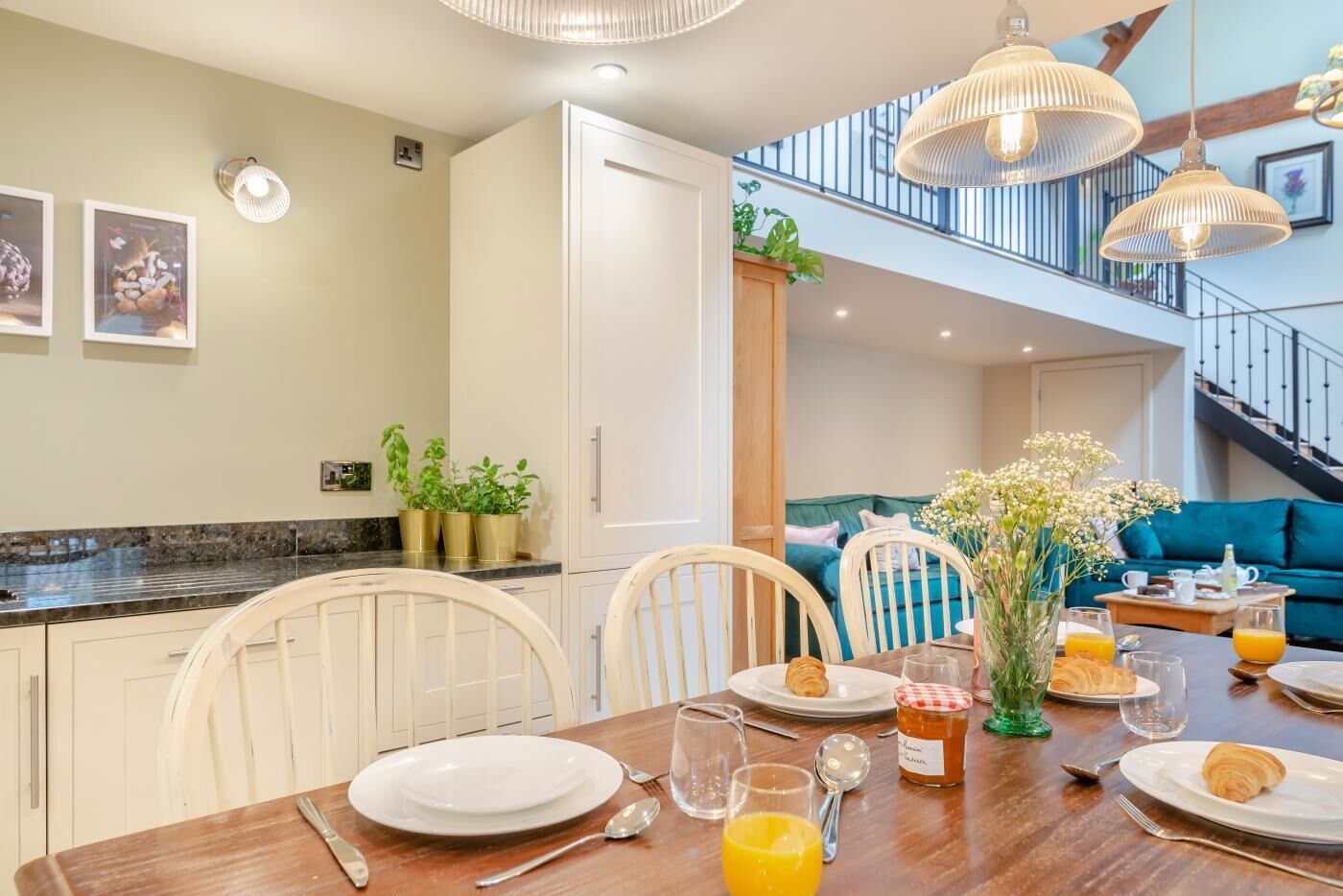 A dining area with a wooden table set for breakfast, including plates, glasses, and a jar of jam. There are potted plants on the counter and a staircase leading to an upper level.