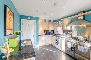 A modern kitchen with a large stove, washing machine, and a dining area with a green table and a bowl of fruit. - 3 Bed in Filey (oc-c30101) (Filey)