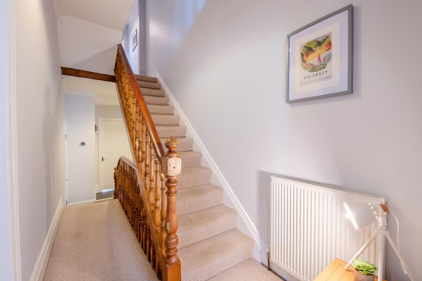 A staircase with wooden balusters and beige carpeted steps leading to a landing. A framed picture hangs on the wall above the landing.