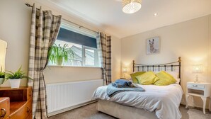 Quarto com cama, janela com cortinas, radiador, mesa de cabeceira com abajur e um quadro na parede.