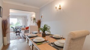 Uma sala de jantar com uma mesa comprida posta para quatro pessoas, um sofá ao fundo e um vaso de planta na mesa.