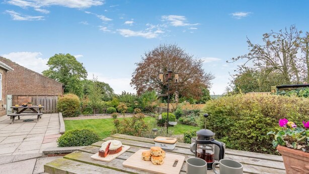 Uma mesa de piquenique de madeira com comida e bebidas, uma casa de tijolos e um jardim com árvores e arbustos.