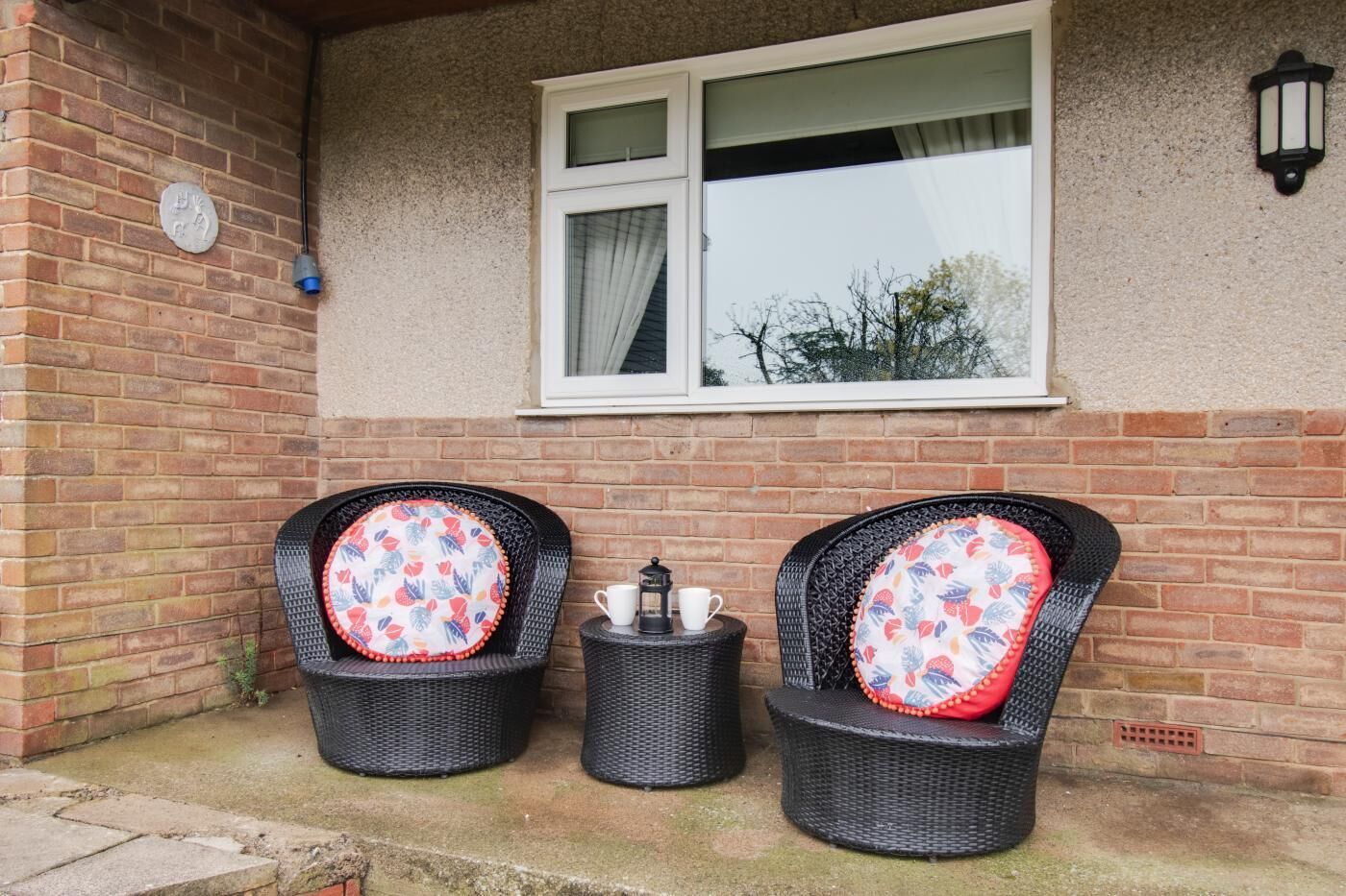 Two wicker chairs with floral cushions, a small table with a lantern, and a window with a curtain.