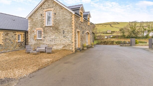 A stone building with a sloped roof, a paved driveway, and a small outdoor seating area with chairs. - 2 Bed in Capel Bangor (oc-m30146) (Capel Bangor)