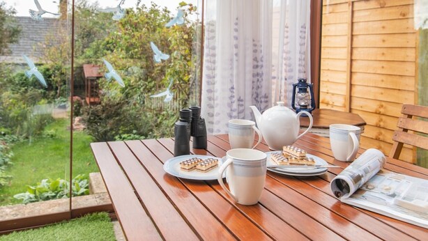 Een houten tafel gedekt met een theepot, kopjes en een bord eten, met uitzicht op een tuin door een glazen raam.