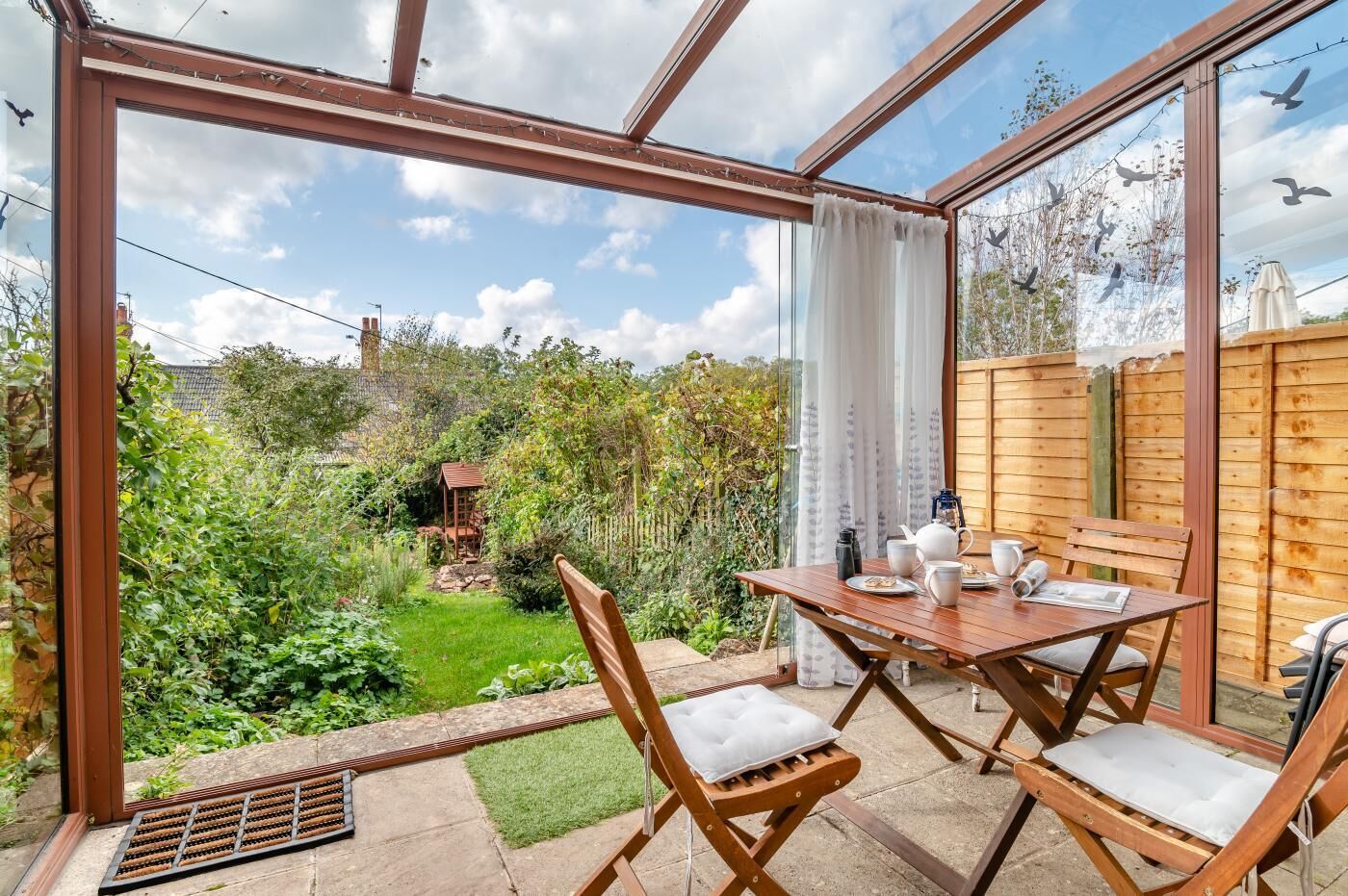 A conservatory with a wooden table and chairs, a garden view, and a wooden fence.