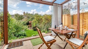 Een oranjerie met een houten tafel en stoelen, uitzicht op een tuin en een houten hek.