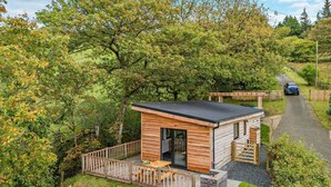 A small wooden cabin with a deck and a car parked on the road in the background. - 1 Bed in Llanerfyl (oc-d30168) (Llanfair Caereinion)