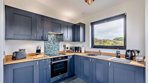 A modern kitchen with dark cabinets, a tiled backsplash, and a window providing a view of greenery. - 1 Bed in Llanerfyl (oc-d30168) (Llanfair Caereinion)