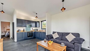 A modern kitchen with dark cabinets, a central island, and pendant lighting. A dark gray sofa with cushions is in the living area, and a wooden coffee table with fruit is in the foreground. - 1 Bed in Llanerfyl (oc-d30168) (Llanfair Caereinion)