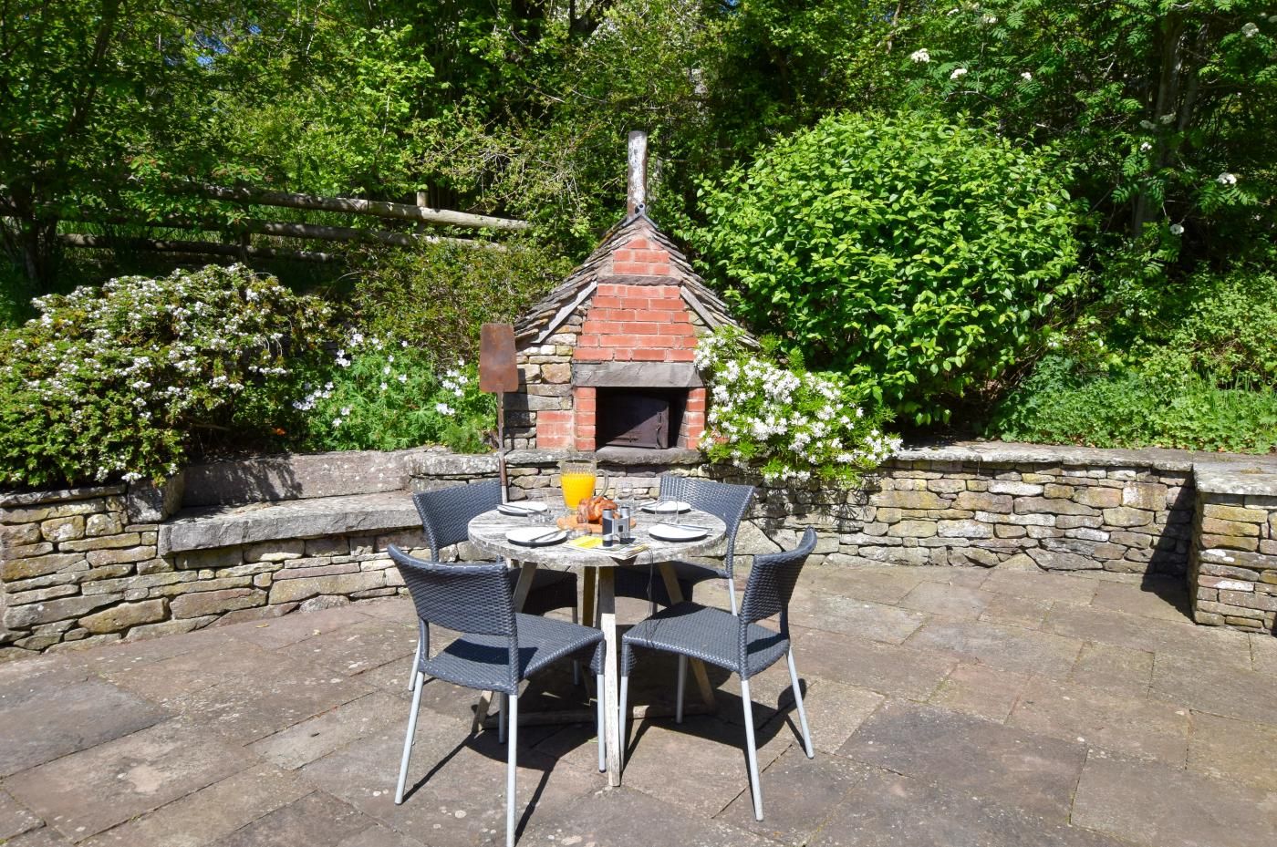 An outdoor dining area with a brick oven, stone walls, and lush greenery.