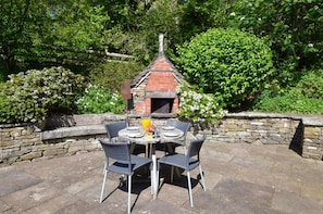 An outdoor dining area with a brick oven, stone walls, and lush greenery.
