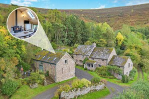 A traditional stone house with a stone staircase leading to the entrance, surrounded by lush greenery and a hilly landscape.
