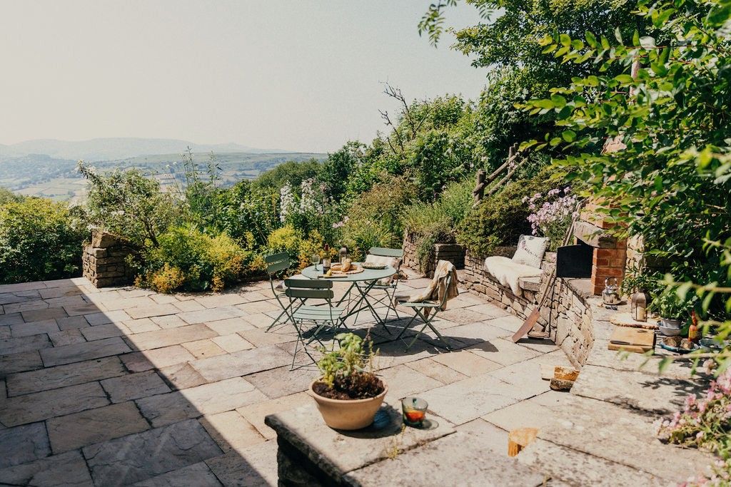 An outdoor patio with stone flooring, a round table, and chairs, surrounded by greenery and a view of distant hills.