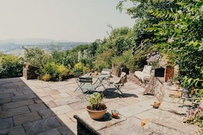 An outdoor patio with stone flooring, a round table, and chairs, surrounded by greenery and a view of distant hills.