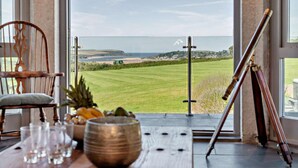 A room with a view of a green field and the sea, a wooden table with glasses and a bowl, and a tripod on the floor. - 5 Bed in St. Issey (oc-r4328) (Saint Issey)