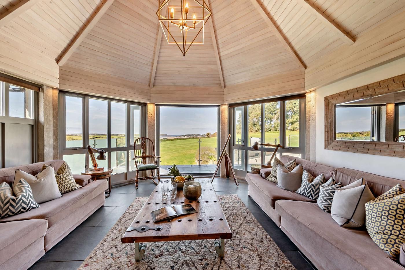 A spacious room with a high ceiling, large windows offering a view of a green landscape, and a wooden coffee table with various items.