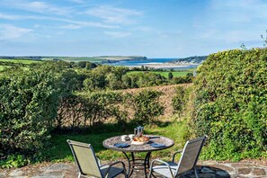 A table with two chairs set outdoors, overlooking a coastal landscape with hills and a beach. - 5 Bed in St. Issey (oc-r4328) (Saint Issey)