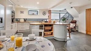 A modern kitchen with white cabinetry, a central island, and a dining area with a table set for breakfast. - 3 Bed in Heacham (oc-t30182) (Heacham)