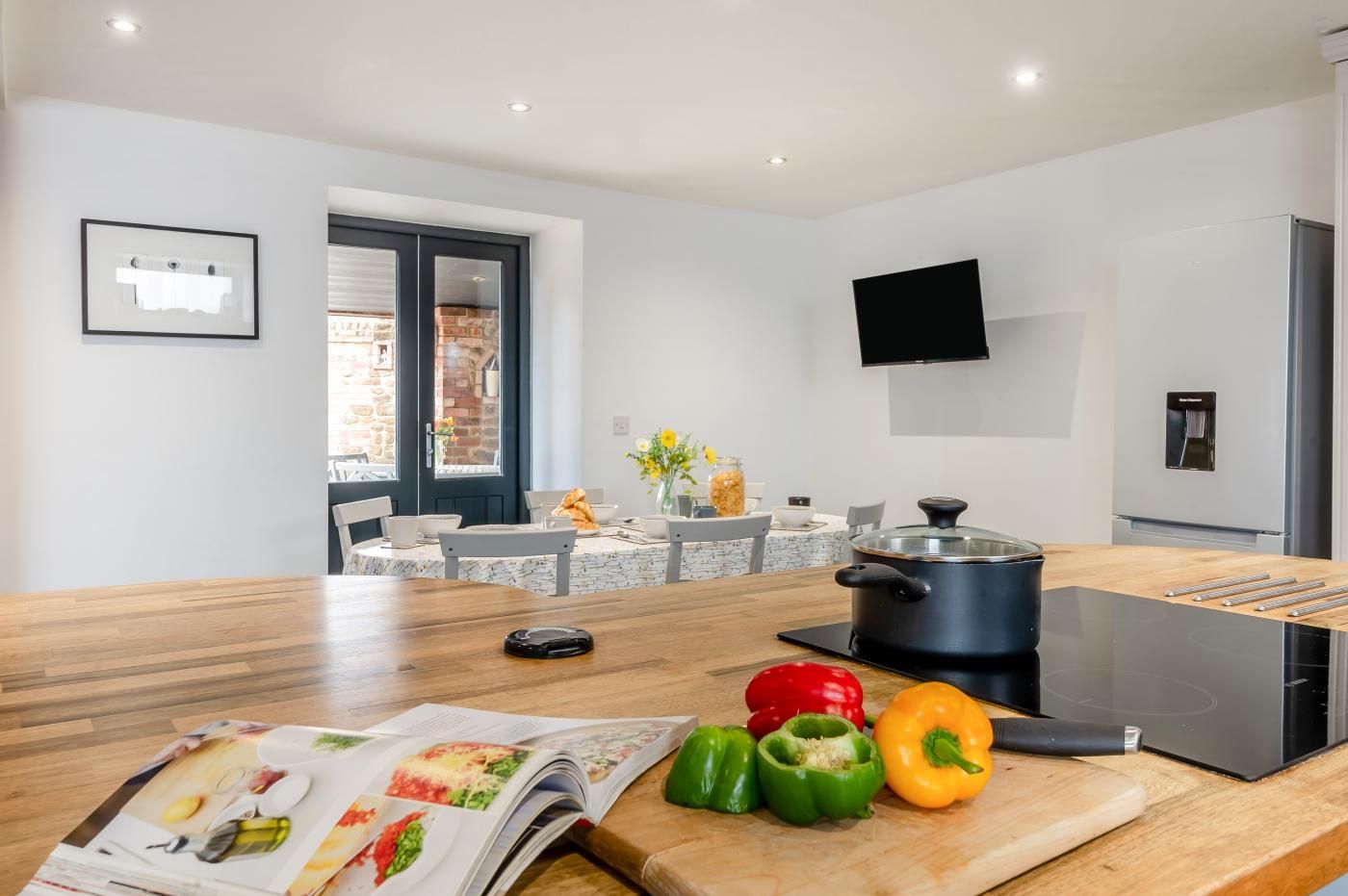 Une cuisine moderne avec un plan de travail en bois, une plaque de cuisson à induction noire et une casserole. On peut voir des légumes sur le plan de travail et un magazine ouvert.
