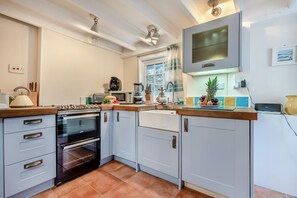 Een kleine keuken met een kookplaat, oven, magnetron en spoelbak. Er is een raam met gordijnen, een klein aanrecht met een waterkoker en een fruitschaal.