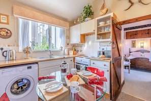 A compact kitchen with a washing machine, a dining area with a glass table, and a bedroom with a bed and a nightstand. - 1 Bed in Ilfracombe (oc-c30176) (Ilfracombe)