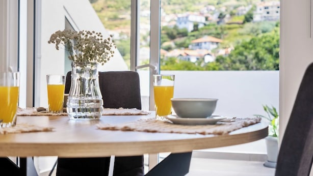 A dining table with a vase of flowers, two glasses of orange juice, and a bowl on a table with a view of a green landscape through large windows. - Apartment Lurdes (Arco da Calheta)