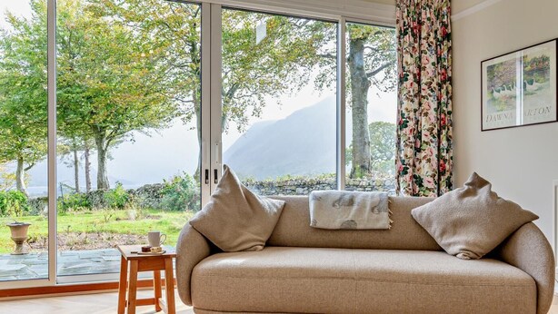 Una sala de estar acogedora con un sofá beige, una pequeña mesa de madera con una taza, y vistas a un jardín exuberante y montañas a través de amplias ventanas.