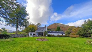 Una casa con un jardín grande, un muro de piedra y una montaña al fondo.