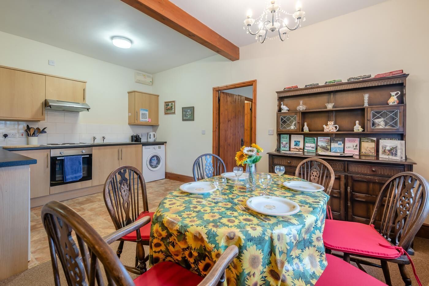A dining area with a round table set for four, surrounded by wooden chairs with red cushions. A chandelier hangs from the ceiling, and a bookshelf filled with books and decorative items is to the right.