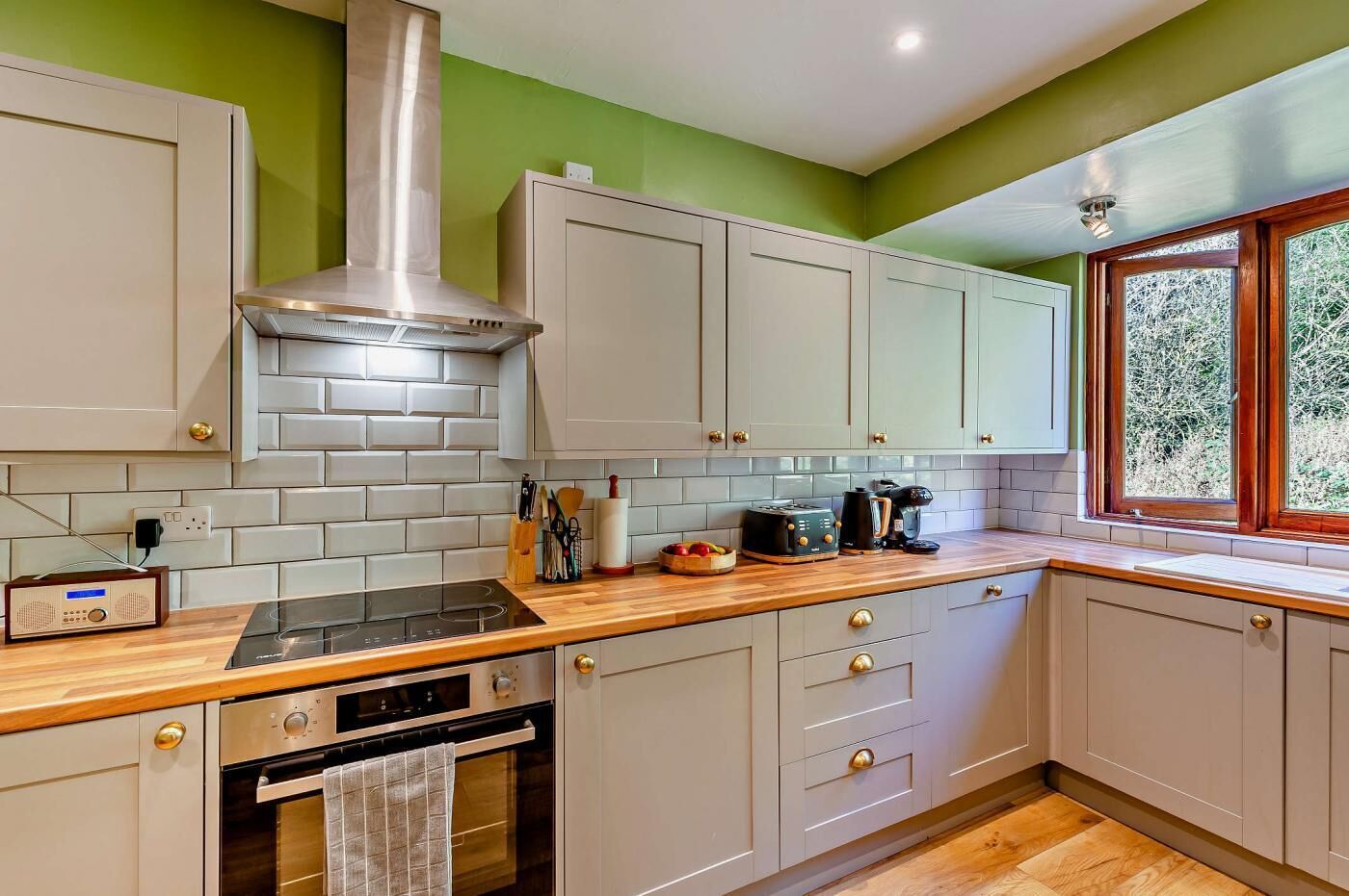 A kitchen with grey cabinets, a stainless steel range hood, a wooden countertop, and a window with a view of trees.