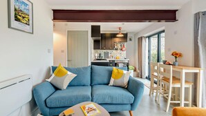 A modern living room with a blue sofa, a dining area with a wooden table and chairs, and a kitchen in the background. - 3 Bed in Ashover (oc-w30175) (Ashover)