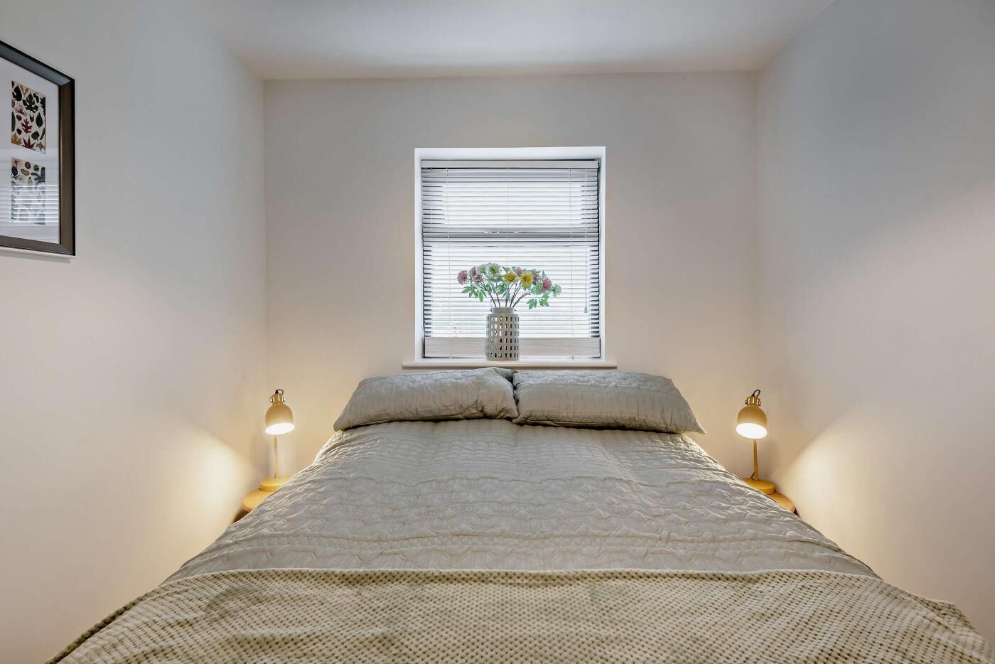 A bedroom with a bed, two bedside lamps, a window with blinds, and a vase with flowers.
