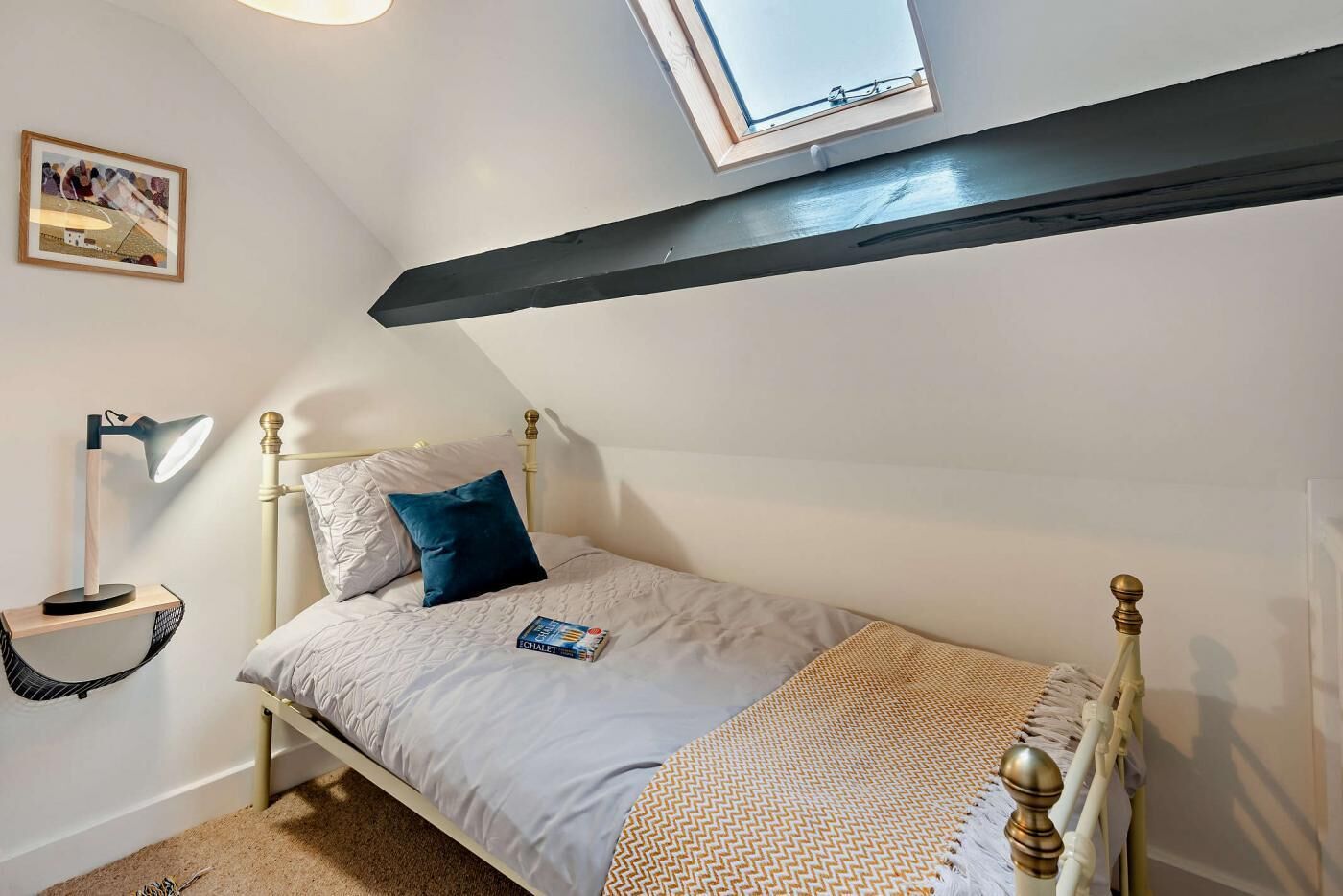 A bedroom with a bed, a bedside lamp, a small shelf, a framed picture, and a skylight.