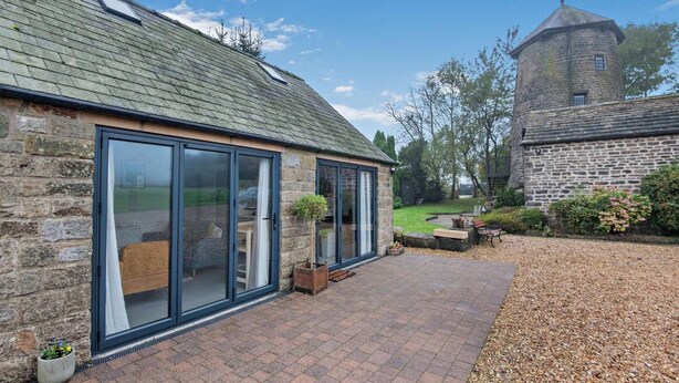 A stone building with large glass doors, a brick patio, and a stone tower in the background. - 3 Bed in Ashover (oc-w30175) (Ashover)