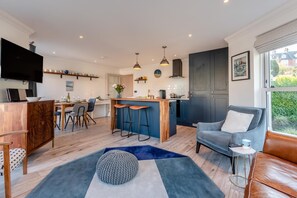 Eine moderne Küche mit einer zentralen Kochinsel, Barhocker und Pendelleuchten. Es gibt eine Essecke mit Tisch und Stühlen, eine Couch und einen an der Wand befestigten Fernseher.
