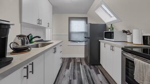 Een compacte keuken met witte kastjes, een magnetron, een waterkoker en een raam met jaloezieën.