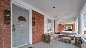 Een overdekt terras met rieten meubels, een grijze deur en een bakstenen muur.