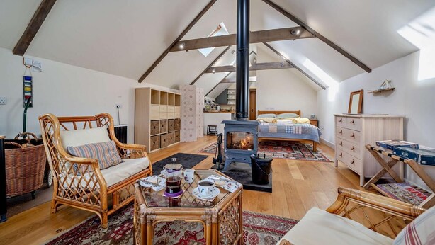 Een ruime loft slaapkamer met een schuine kap, een ingebouwde houtkachel, een grote kledingkast en een gezellige zithoek met rieten meubels.
