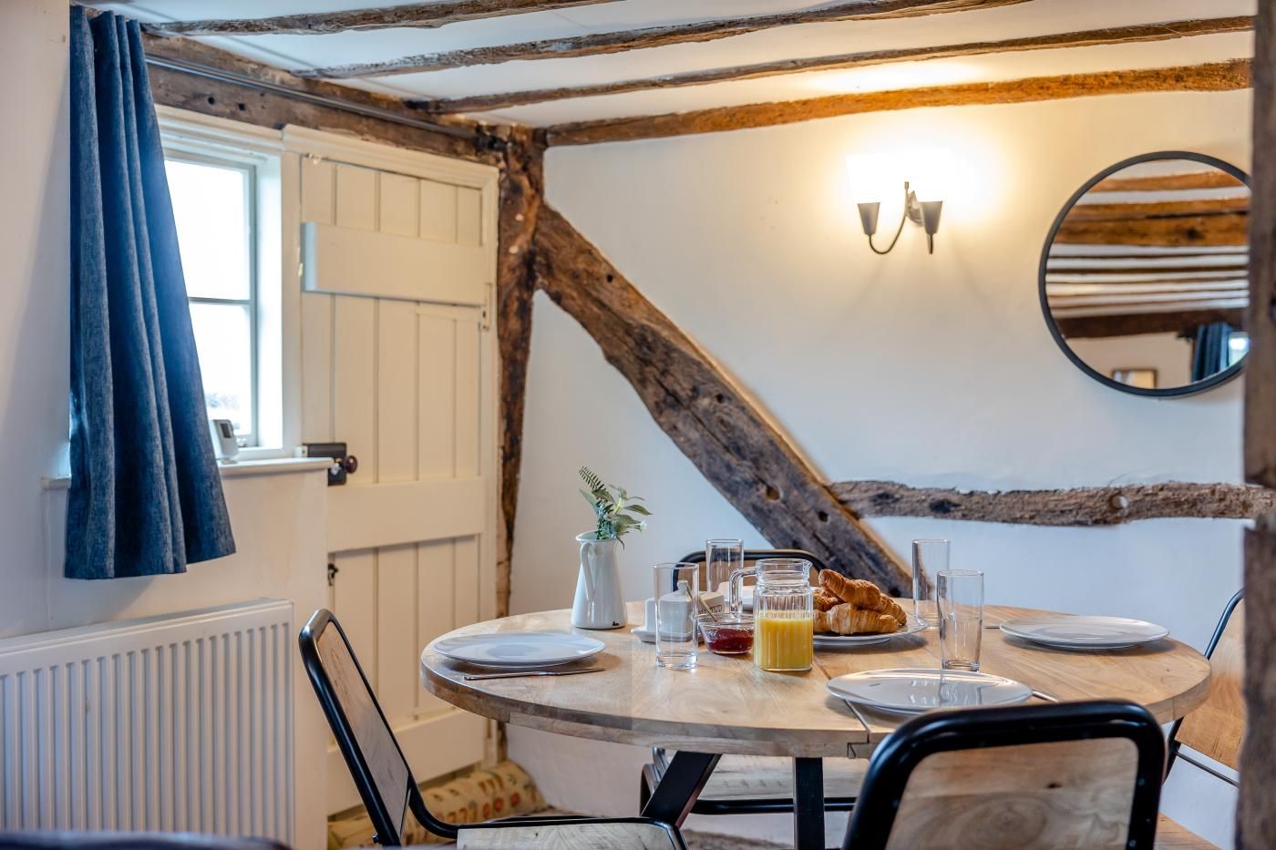 A dining area with a round table, chairs, a window with curtains, a wall-mounted light fixture, and a rustic ceiling beam.