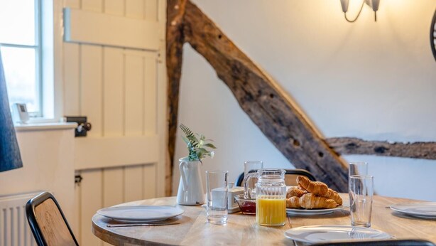 Ein Esstisch mit Croissants, Saft und einem Krug, in einem Zimmer mit sichtbarem, rustikalen Holzbalken und Blick ins Freie.
