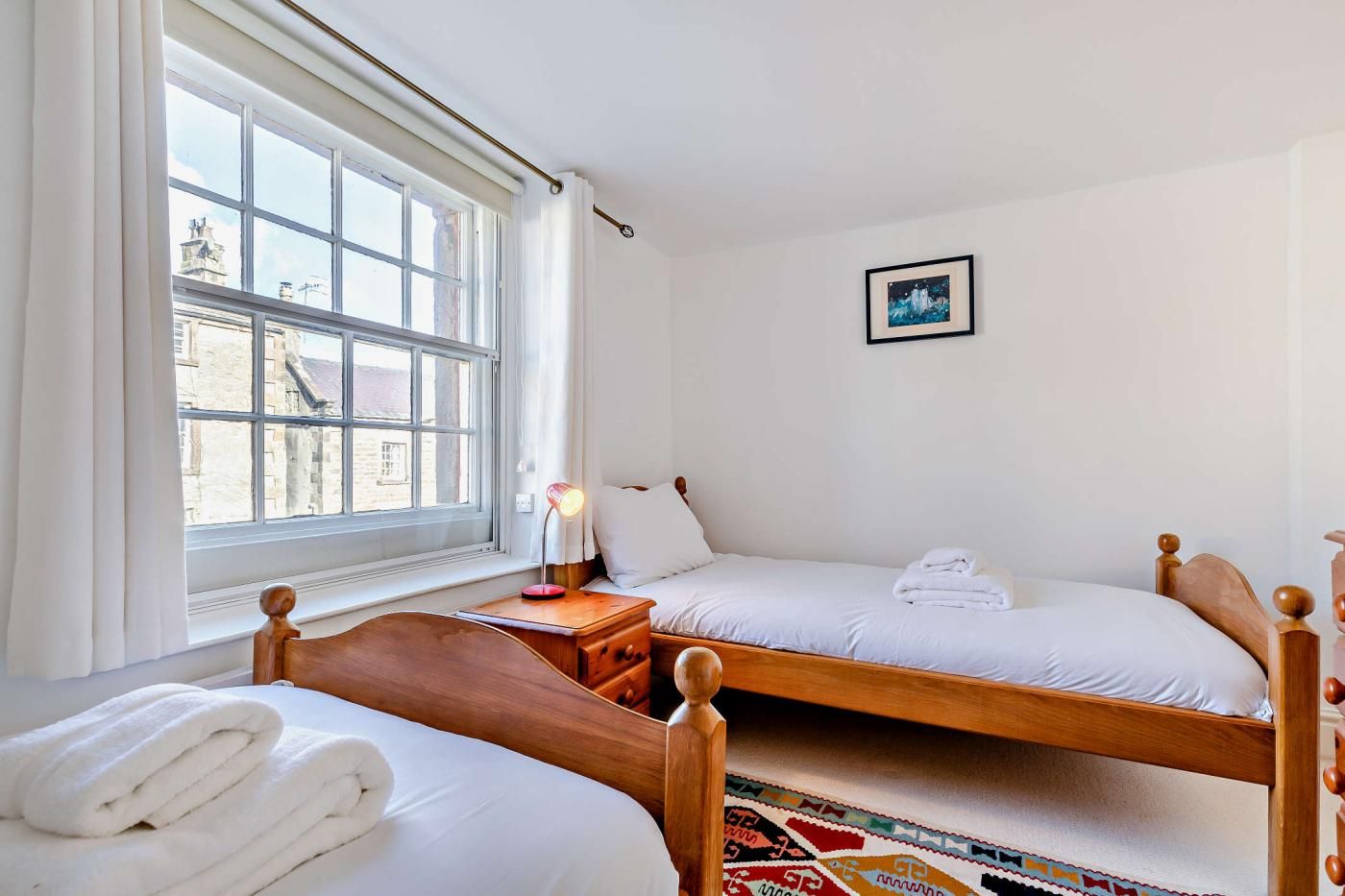 A bedroom with two beds, a window with curtains, a bedside table with a lamp, and a framed picture on the wall.