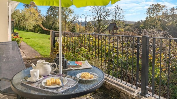 Taula rodona de vidre amb una cadira metàl·lica, un para-sol verd, una càmera, una tetera, tasses i un plat de pastissos en un balcó amb vista a un jardí i arbres.