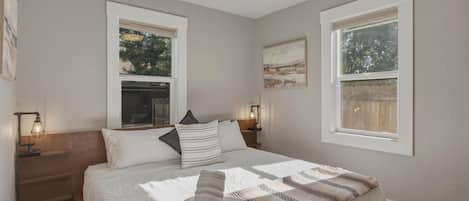 A bedroom with a large bed, two bedside tables with lamps, a window with a view of trees, and a painting on the wall.