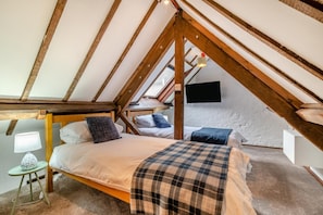 Une chambre avec un plafond en pente, des poutres en bois, un lit, une table de chevet avec une lampe et un téléviseur fixé au mur.