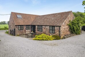 A stone farmhouse with a gravel driveway, a small outdoor seating area, and a fenced property.