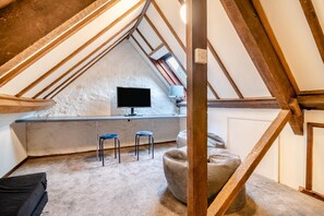 A loft bedroom with a sloped ceiling, a television, two stools, and a window.