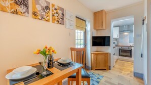 Un comedor con una mesa de madera dispuesta para dos personas, un jarrón de tulipanes y una botella de champán. Se observan pinturas florales enmarcadas en la pared. Se ve una cocina al fondo con microondas, horno y armarios de madera.