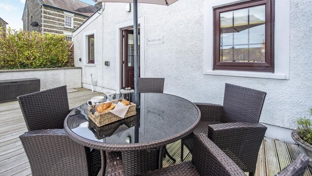 Un espacio de comedor al aire libre con sillas de mimbre y una mesa de vidrio, un edificio blanco con una ventana, y un seto al fondo.